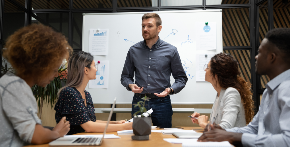 A team of professionals in a meeting around a table, listening to a colleague presenting information on a whiteboard. The charts and notes on the board illustrate data and ideas, while the participants take notes and use laptops to collaborate. The atmosphere is professional and collaborative, reflecting a dynamic and solution-focused work environment.